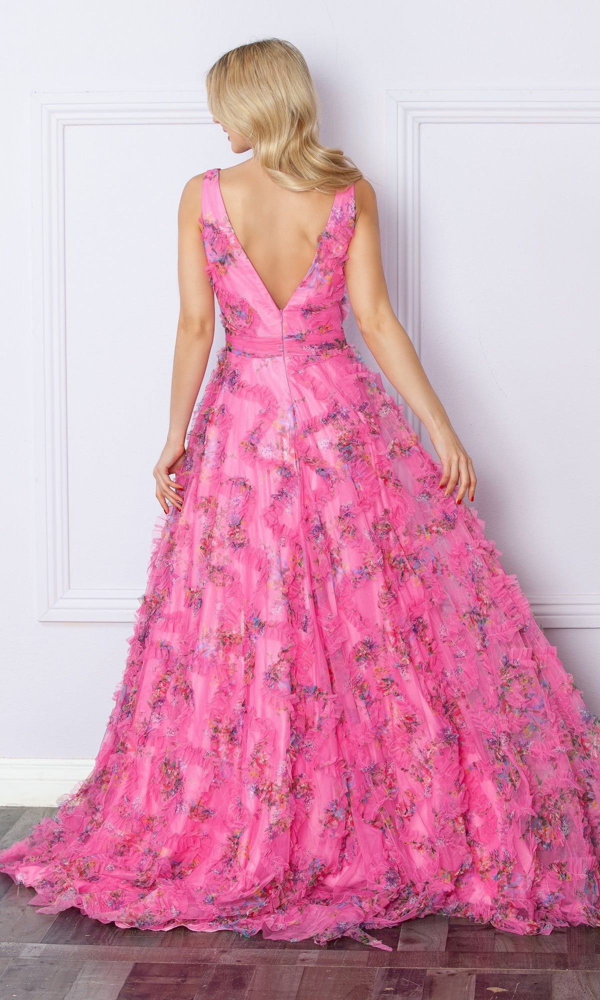 A woman with blonde hair wears the Nox Anabel E1445 Pastel Print Long Prom Dress, a sleeveless, floor-length pink floral gown with a deep V-back, standing against a light-colored wall and facing away from the camera.